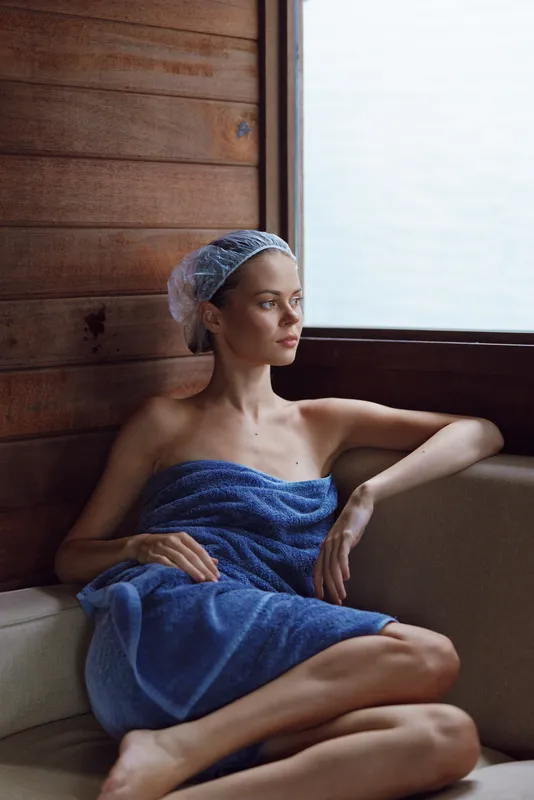 Relaxed woman wrapped in a blue towel and hair cap sitting indoors by wooden wall near window, enjoying calm and peaceful spa or sauna atmosphere.