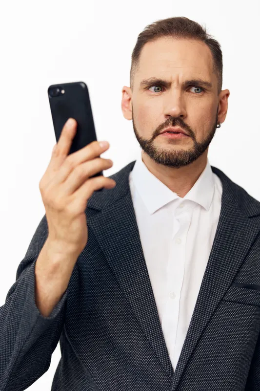 A professional man in a blazer and white shirt looks at his phone with a focused, puzzled expression, photographed against an isolated colored background.