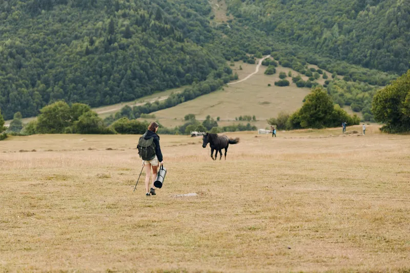 A lone traveler walks across a wide grassy field toward distant hills. Carrying a backpack and hiking gear, they explore a tranquil rural landscape.