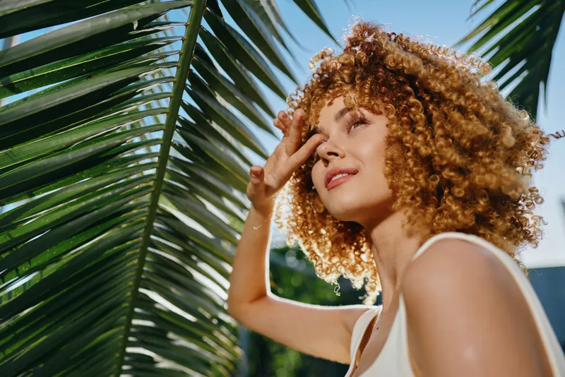 Curly woman shielding eyes from sunlight near tropical palm leaves, enjoying outdoors with warm summer vibes in bright natural light.