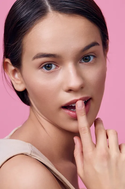 A young woman with a charming smile and finger on lips against a pink backdrop, conveying a sense of playful secrecy and confidence, representing beauty concepts