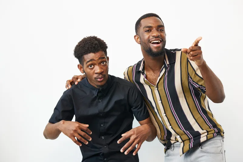 Two cheerful black men enjoying a playful moment together, showcasing friendship and joy in their vibrant clothing against a light background.