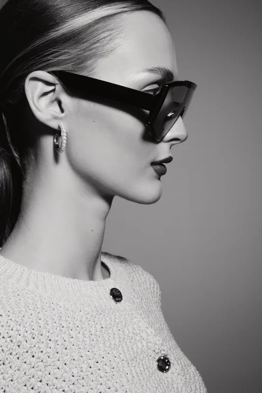 Profile of a Caucasian woman with pale skin wearing dark sunglasses and hoop earrings in a monochrome studio portrait.