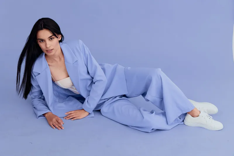 Young woman in blue suit reclining on matching blue floor and background, wearing white sneakers and beige top, with long dark hair, showcasing modern fashion style.