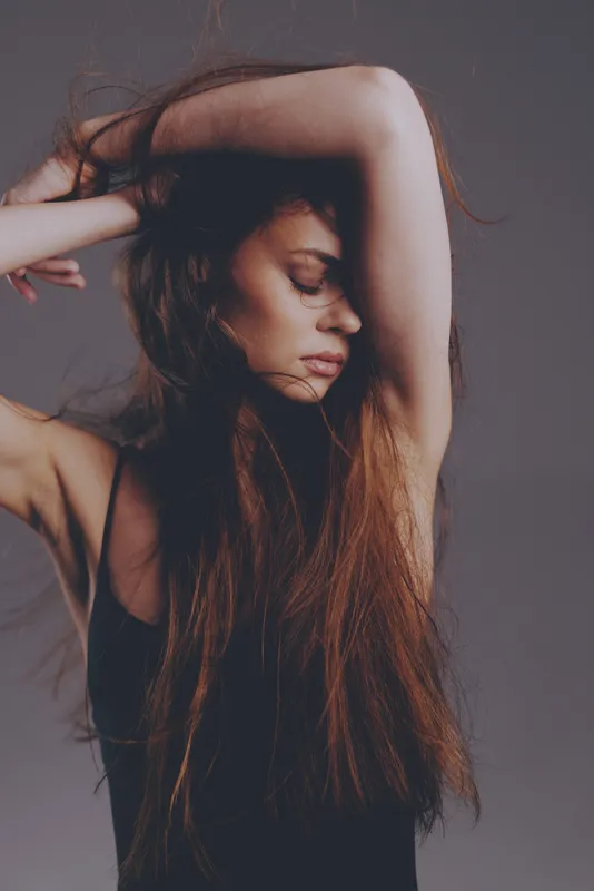 Young woman with long hair posing thoughtfully with arms raised, expressing calmness and introspection against a neutral background