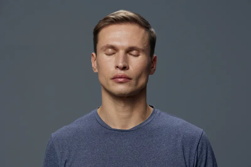Calm young man with closed eyes and relaxed expression wears casual blue sweater against neutral gray background. Studio portrait capturing serenity, mindfulness, and tranquility in a minimalist style