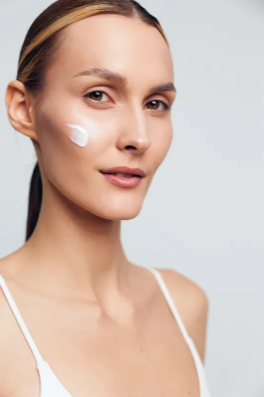 Young woman applies skincare cream to her cheek in a close-up studio portrait with soft lighting and a clean white background, conveying calm confidence and focus on self-care and healthy skin.