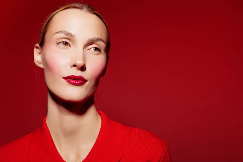Portrait of a confident woman with red lipstick and red jacket on red background. Studio lighting highlights her clear skin and elegant expression. Fashion and beauty concept.