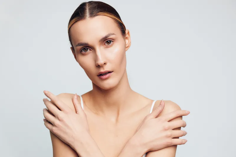 Portrait of a young woman with clear skin and natural makeup posing with crossed arms on a light background. Concept of beauty, skincare, and minimalism with soft studio lighting.