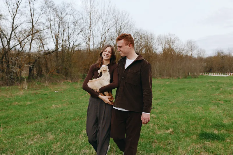 Smiling couple strolling outdoors with a goat kid in a field, under soft sunlight. This lifestyle scene conveys affection, companionship, and rustic charm in a natural setting.
