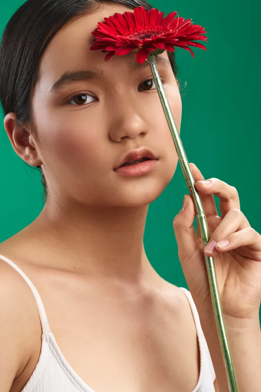 Asian woman is seen holding a flower at close range in a simple studio setting. She gazes softly at the camera, conveying calm beauty and a serene, intimate mood.