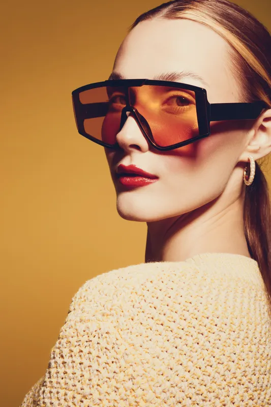 Stylish woman with oversized orange sunglasses and red lipstick poses against yellow background. Fashion portrait with warm lighting and trendy accessories for modern look.