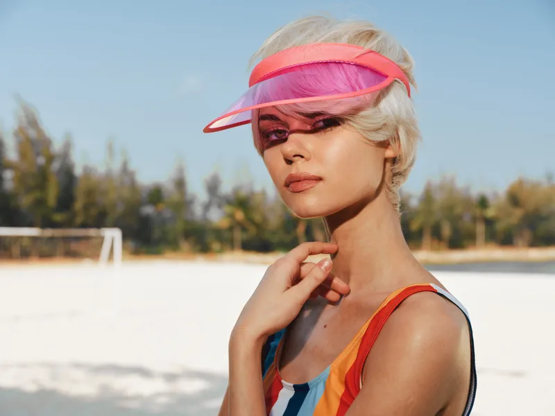 A stylish woman stands on a sunlit beach wearing a vibrant striped swimsuit and a pink visor, looking into the distance with calm confidence as wind lifts her hair.