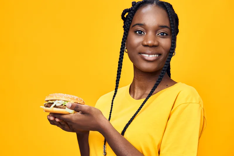 Smiling African woman holding a delicious burger against a vibrant yellow background, symbolizing healthy eating and joyful dining experiences