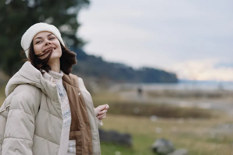 woman outdoor winter jacket beanie smiling in a breezy coastal landscape with soft light and calm mood, portraying warmth and comfort in nature