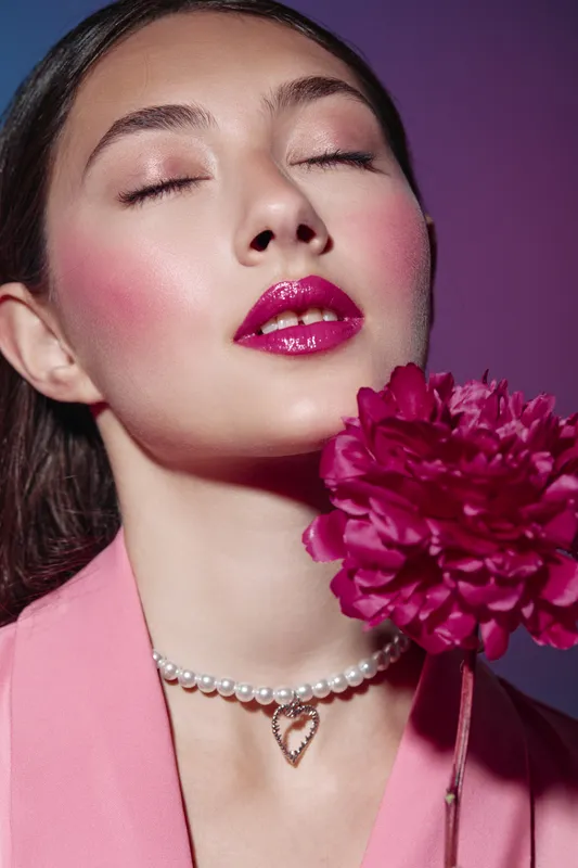 Close-up of a young woman with glossy fuchsia lips and pink blush. She wears a pearl necklace and pink blazer while holding a matching vibrant flower. Fashion beauty concept.