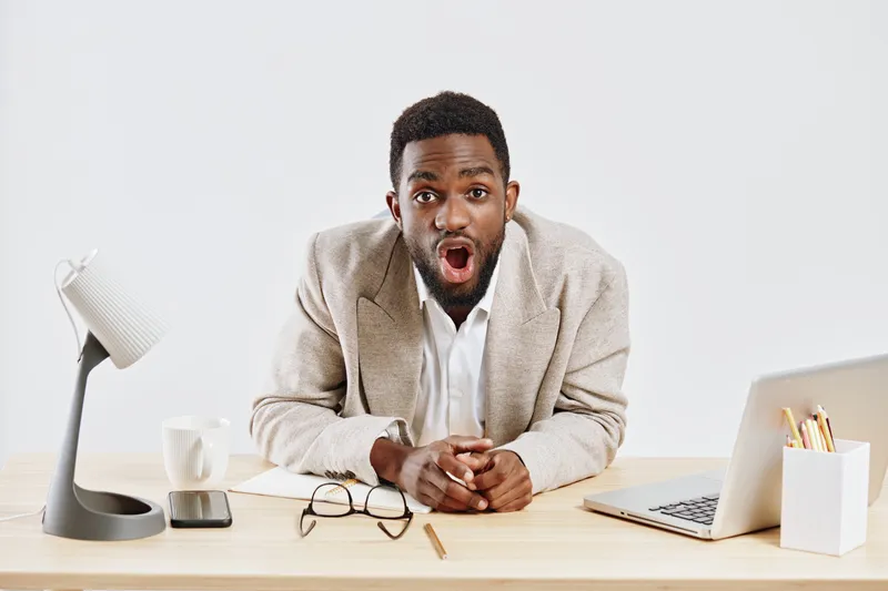 Excited young man with dark skin sitting at office desk looking surprised and engaged, wearing beige blazer and white shirt, with laptop, eyeglasses and stationery on plain white background. Business 