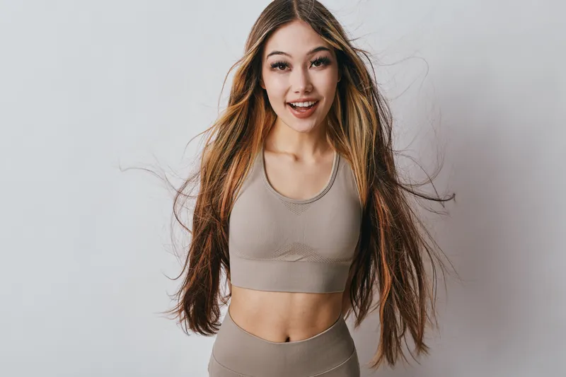 Young woman with long hair wearing sports bra and leggings smiling in studio for fitness, wellness and healthy lifestyle concept with white background and natural look.
