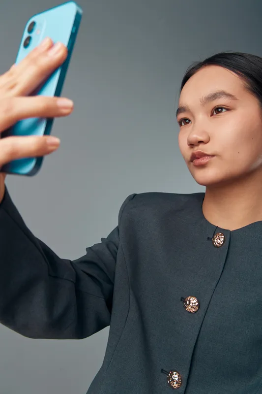 Young woman taking selfie with smartphone in studio. Portrait of female with serious expression wearing dark blazer with gold buttons on gray background.