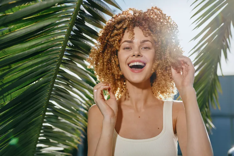 Smiling woman with curly hair enjoys sunny day outdoors near green palm leaves, wearing white tank top and expressing joyful summer mood.
