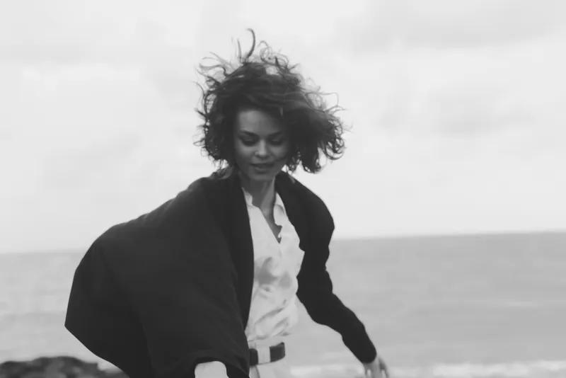 Black and white photo of a woman on the coast, hair blown by wind, wearing a jacket and white shirt, capturing motion and coastal style