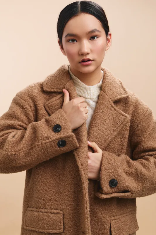Asian woman wearing a warm brown coat stands in a neutral studio, looking directly at the camera with a calm, confident expression that conveys style and poise.
