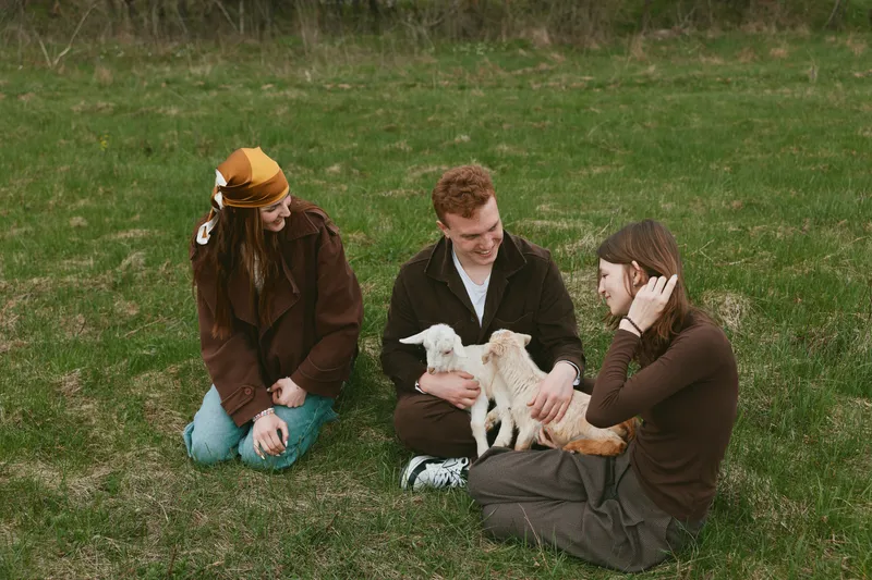 Goat, kid, people, outdoors, friends, interaction captured as a trio enjoying gentle petting on a grassy field, rustic clothing, calm mood, natural light, candid rural lifestyle moment, warm
