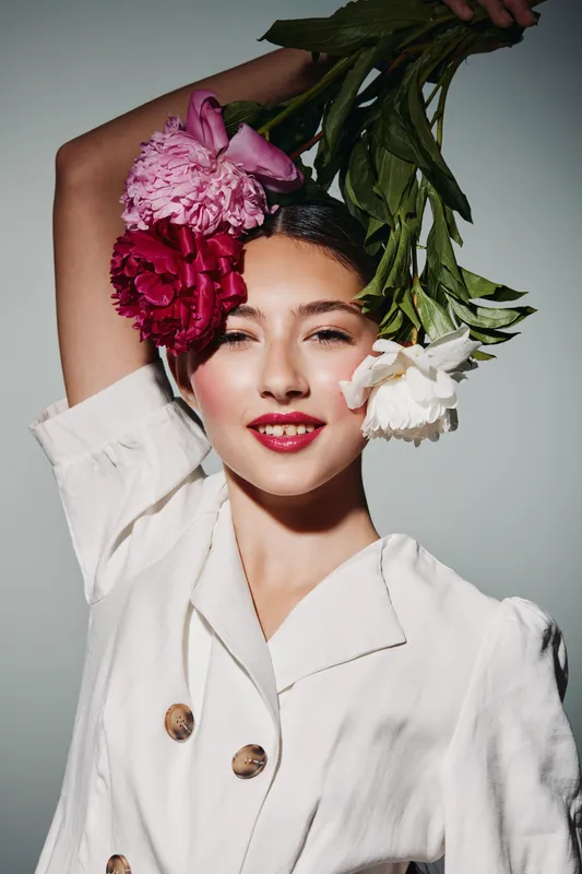 Young woman in white blazer holds vibrantly colored flowers above her head, smiling with bold red lipstick. Bright studio light highlights beauty, fashion, and creative floral styling.