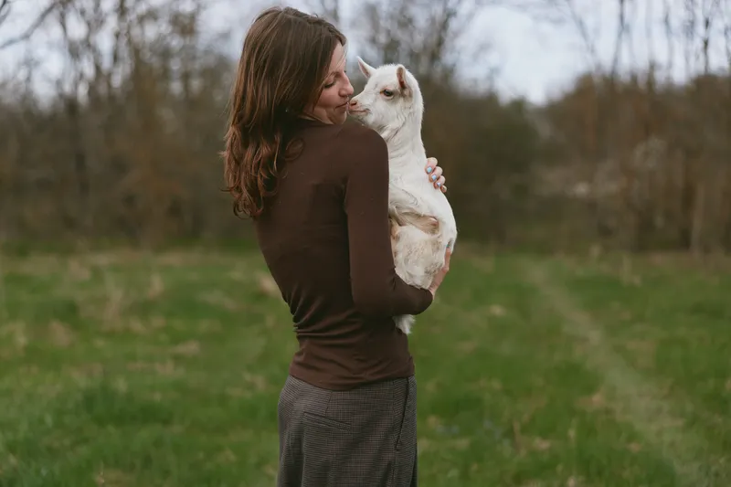 A woman in a brown sweater holds a young goat in an open field, sharing a calm moment of care and connection with nature. A lifestyle scene emphasizing animal companionship and gentle, everyday