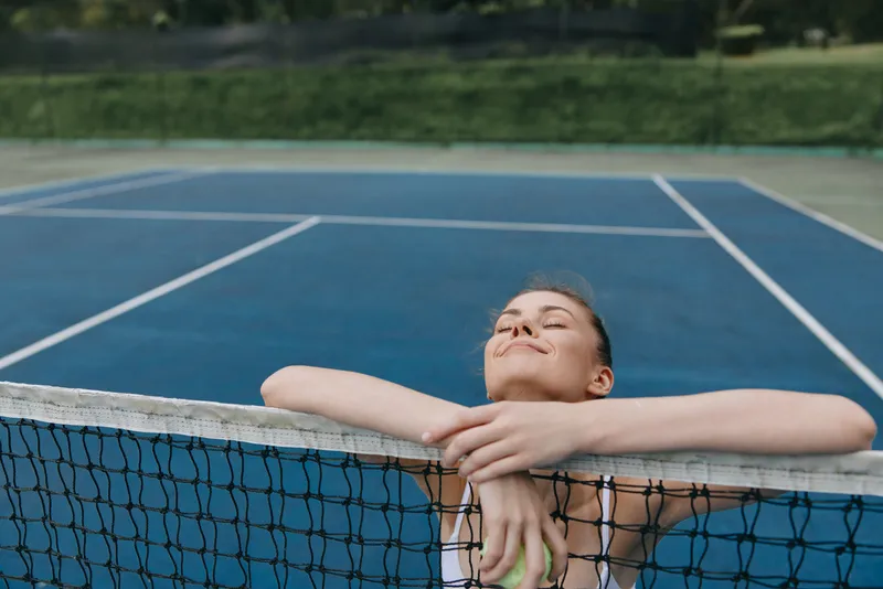 Young woman in sports attire relaxing by a tennis net, enjoying a break on a blue court, radiating happiness and energy, capturing the essence of active lifestyle