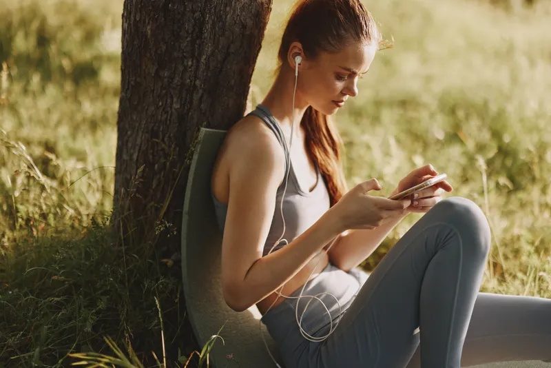 Young woman enjoying music on her smartphone while sitting outdoors in activewear, representing a healthy lifestyle and relaxation in nature