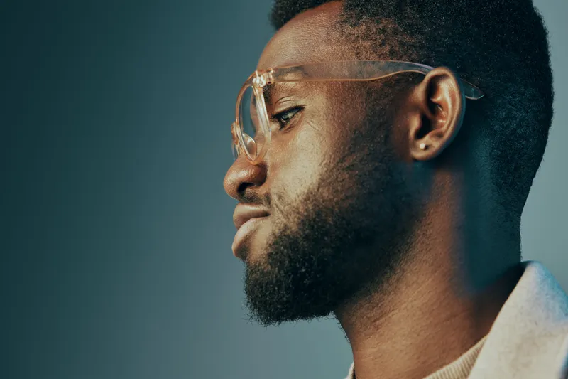 Thoughtful young African man with glasses gazing sideways, showcasing confidence and style against a gradient blue and green background