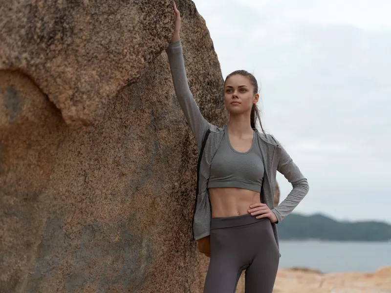 A fit young woman in activewear poses confidently against a rocky background by the sea, embodying strength and determination Fit, woman, activewear, strength, confidence, rocky background