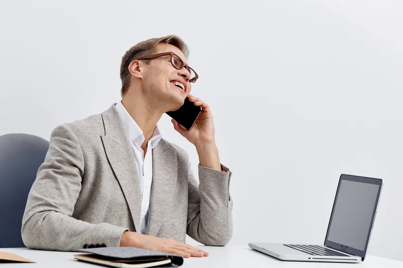 Professional businesswoman wearing glasses and gray blazer smiling while talking on mobile phone, sitting at desk with open laptop, modern office background, corporate communication concept.
