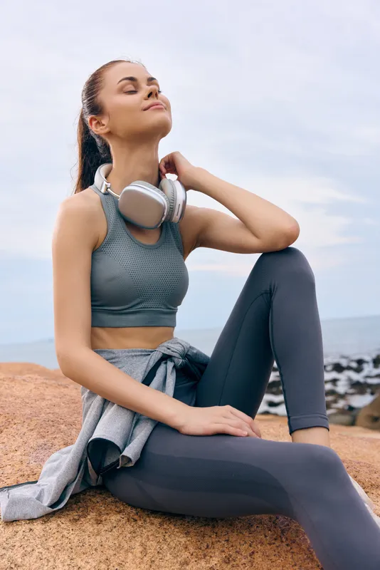 Young woman enjoying outdoor fitness, wearing gray sportswear, headphones, sitting on rocks by the water, exuding calmness and positivity in a natural setting