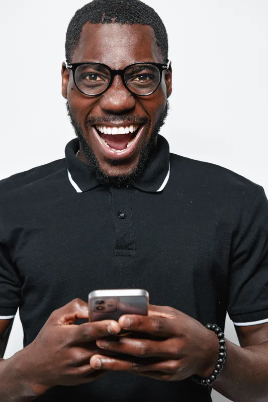 African American man with dark skin wearing glasses and black polo shirt smiling and holding smartphone, happy expression and casual style on white background in studio portrait.