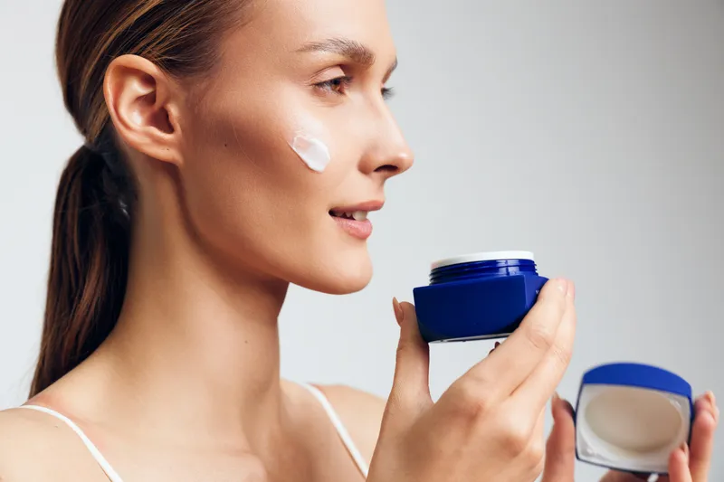 Young woman applies moisturizing cream on her face holds blue jar with skincare product shows gentle skin care routine isolated on white background with natural makeup and serene expression