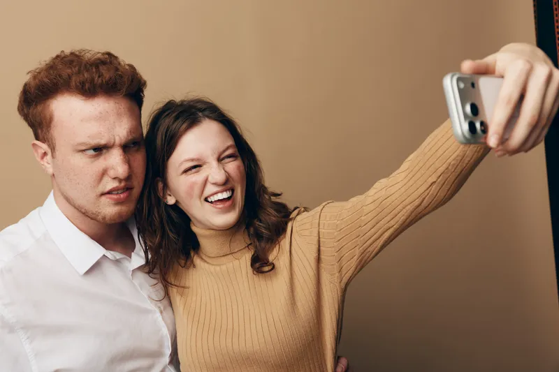 Young couple takes a selfie with a smartphone, woman smiling and man making a confused face. Indoor portrait with casual clothing and neutral background. Concept of relationships and emotions.