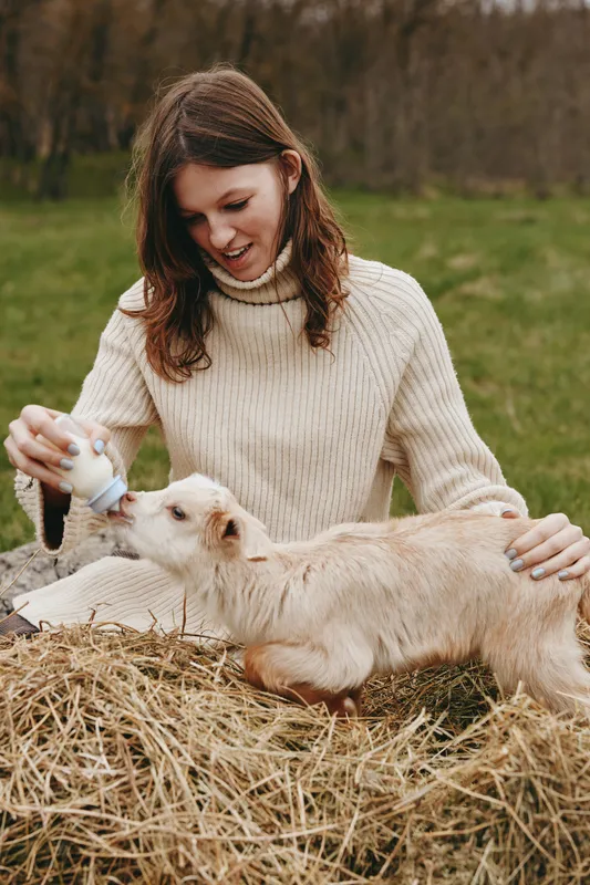 Joyful woman outdoors feeding a goat kid on a hay bale, sunlit field, cozy knit sweater, gentle interaction, soft breeze, natural colors, rural lifestyle moment