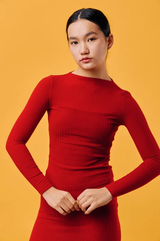 Woman wearing red long sleeve fitted dress standing in front of orange background with hands clasped and straight black hair tied back for fashion portrait.