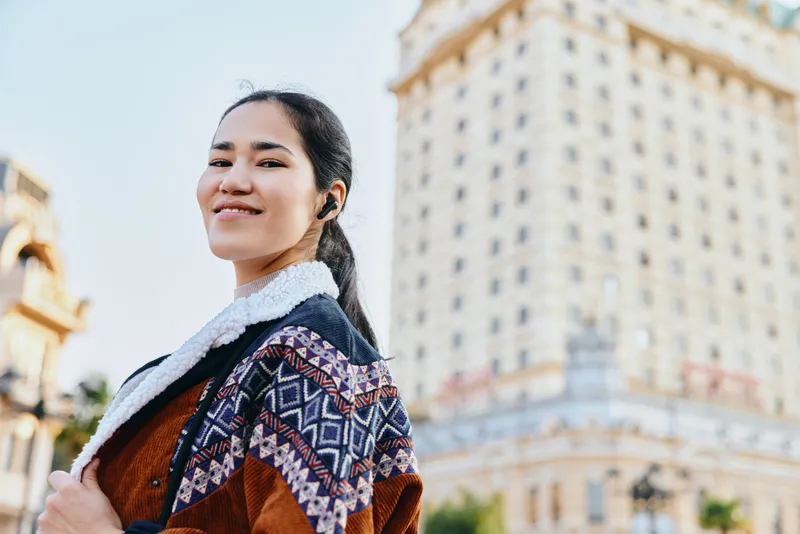 Woman in warm patterned shawl and jacket posing with confident smile against urban cityscape, street style photography showcasing fashion, travel vibe, and sunny daylight in a cosmopolitan setting