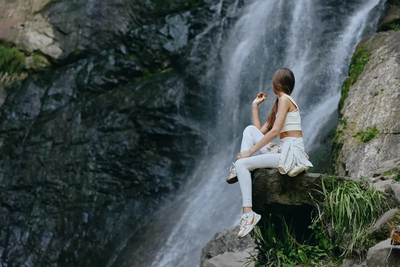 young woman sitting near waterfall in casual white outfit, relaxed, nature, outdoors, adventure, peaceful moment