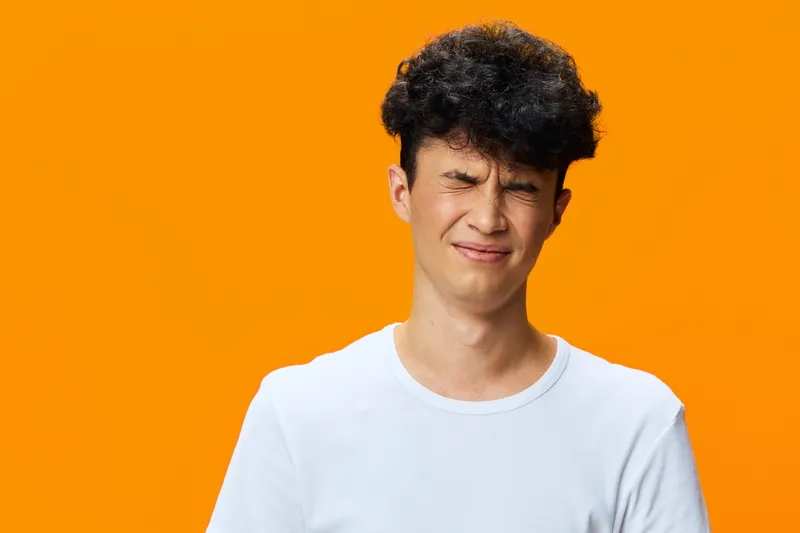 Young man with curly hair wearing a white t-shirt grimacing with eyes closed against bright orange background, expressing discomfort or confusion.