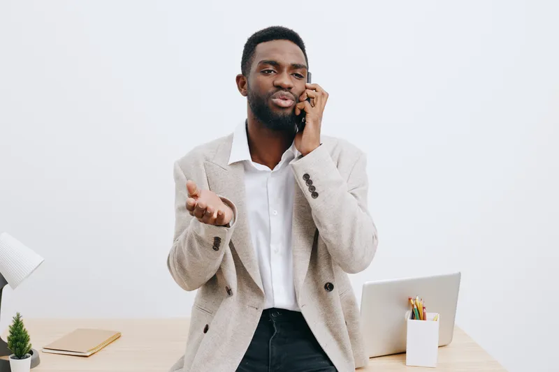 Confident young man talking on mobile phone indoors at modern workspace, wearing smart casual outfit and jacket, expression engaging, professional setting with laptop, stationery, minimal decor, brigh
