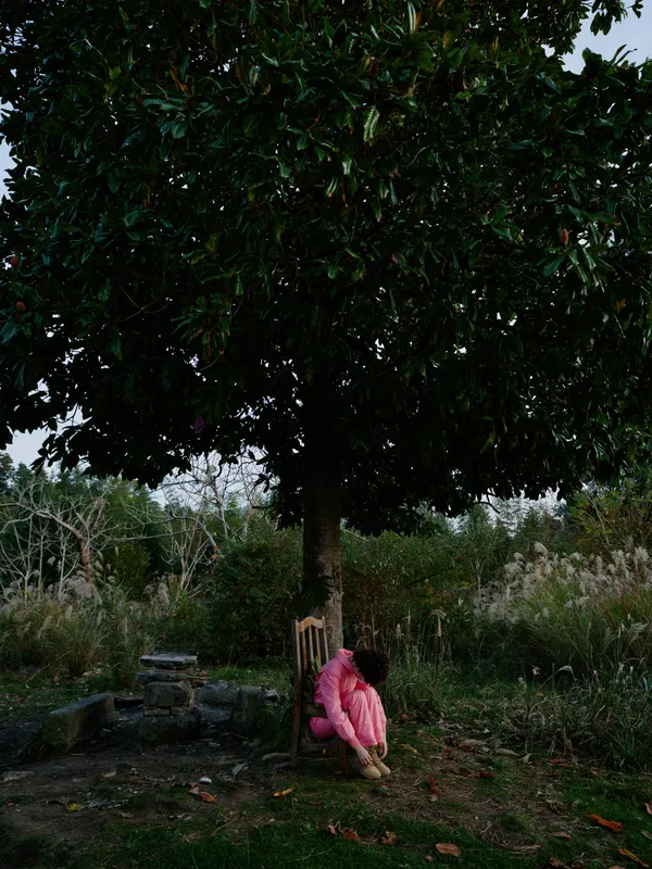 A person in a pink outfit sits under a large tree in a quiet garden, enjoying calm shade and natural scenery. The scene emphasizes solitude, outdoor relaxation, and simple connection with nature in a