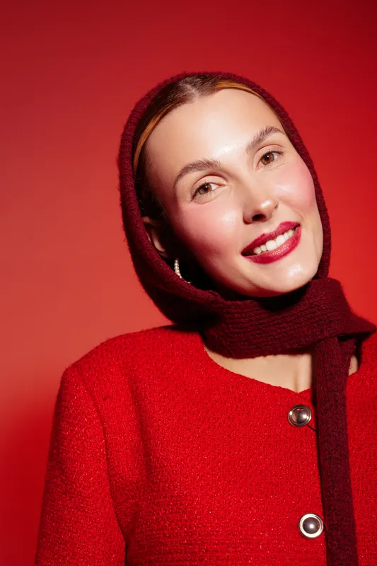 Smiling woman in red coat and scarf poses against red background. Close-up portrait with natural makeup and bright lipstick. Concept of fashion, beauty, and winter style.