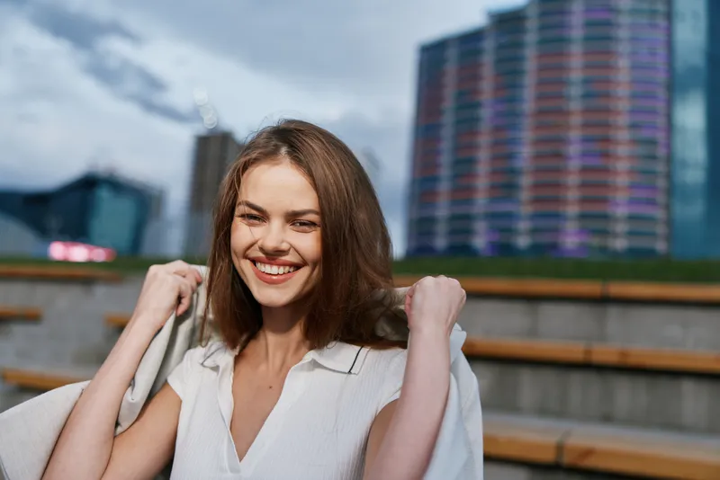 Confident woman in business attire smiling outdoors with modern city background, exuding professionalism and positivity in a dynamic urban setting.