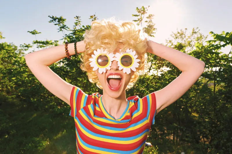 Playful woman outdoors wearing a striped shirt and flowered sunglasses, posing with a wide smile. Bright color palette captures lifestyle, happiness, and carefree summer mood.