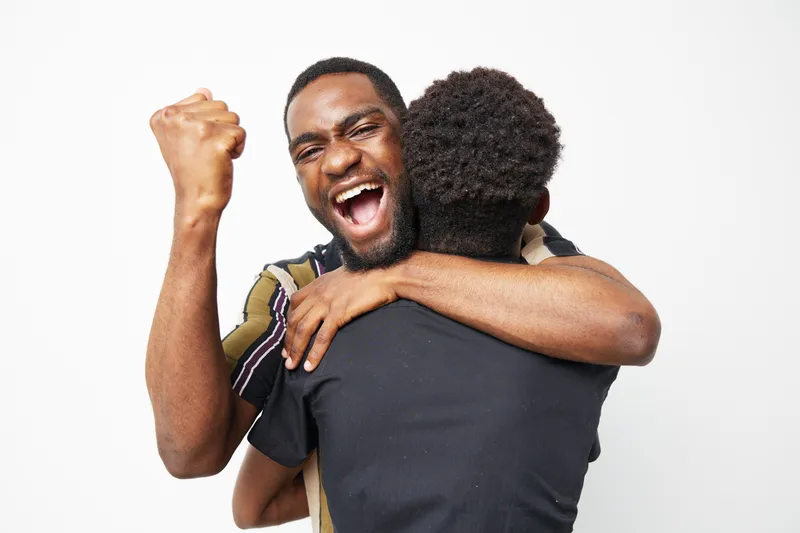 Joyful black men share an exuberant embrace, capturing a moment of celebration and triumph with vibrant energy and camaraderie in a minimal backdrop.