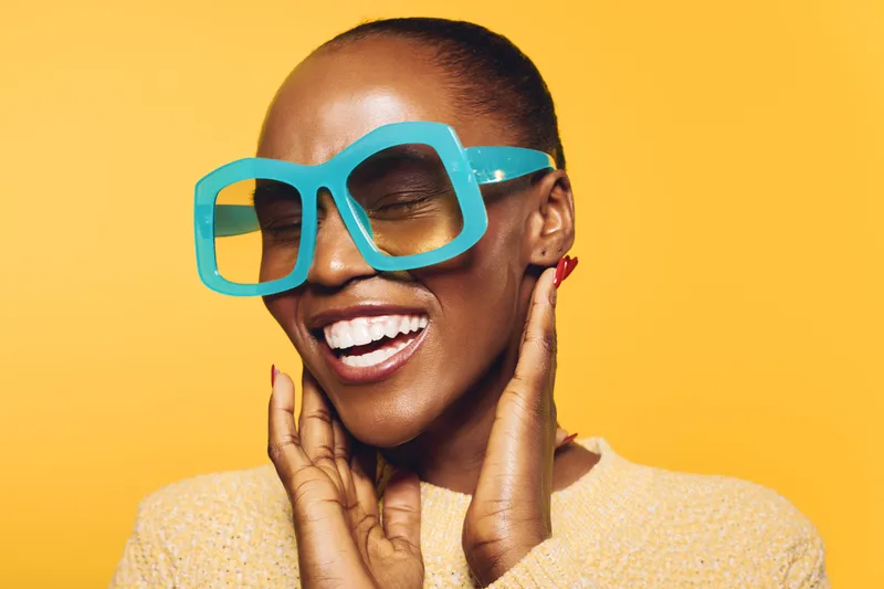 Happy African woman with dark skin wears large blue glasses and a beige sweater, smiling with eyes closed against yellow background. Concept of joy, fashion, and beauty.